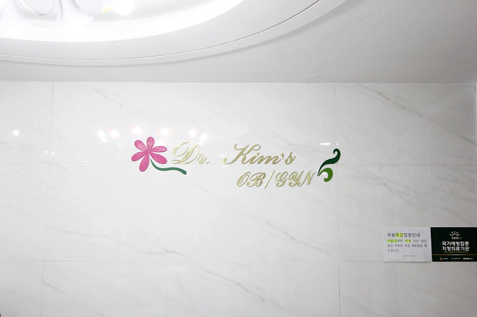병원 로비 대리석 벽면에 부착된 'Dr. Kim's OB/GYN' 금색 로고와 꽃 장식, 우측 하단 독감 예방접종 안내문