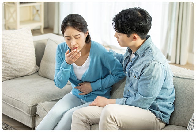 소파에 앉아 복부를 감싸며 아파하는 여성과 옆에서 어깨를 잡아주는 남성을 담은 연출용 스톡사진