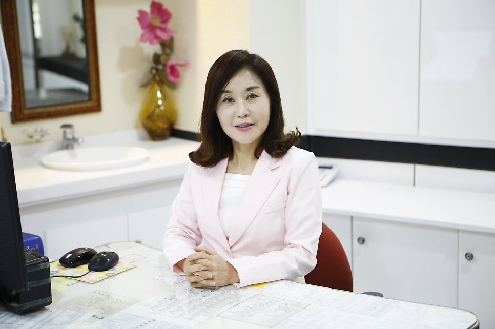 핑크색 재킷을 입은 중년 여성이 진료실 책상 앞에 앉아 손을 모으고 정면을 바라보는 사진